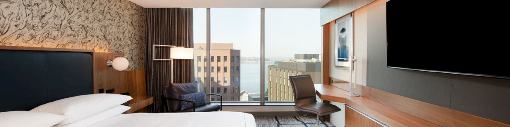 A modern hotel room with a king-size bed, large TV, desk, chair, and a view of buildings outside through a large window.