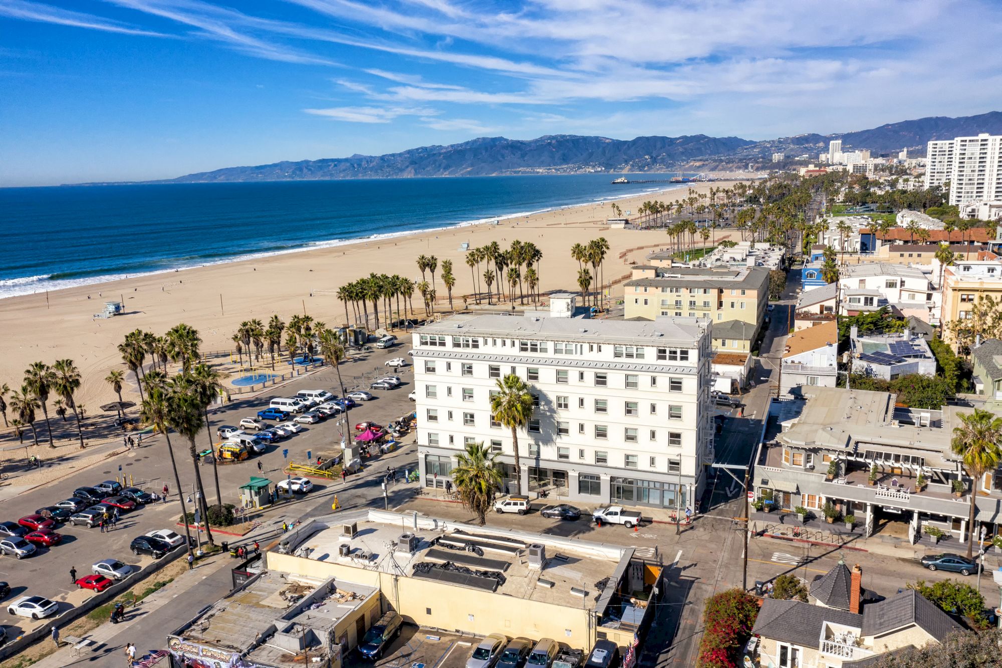 Coastal cityscape with a beach, palm trees, parked cars, and buildings next to the ocean under a partly cloudy sky ending the sentence.