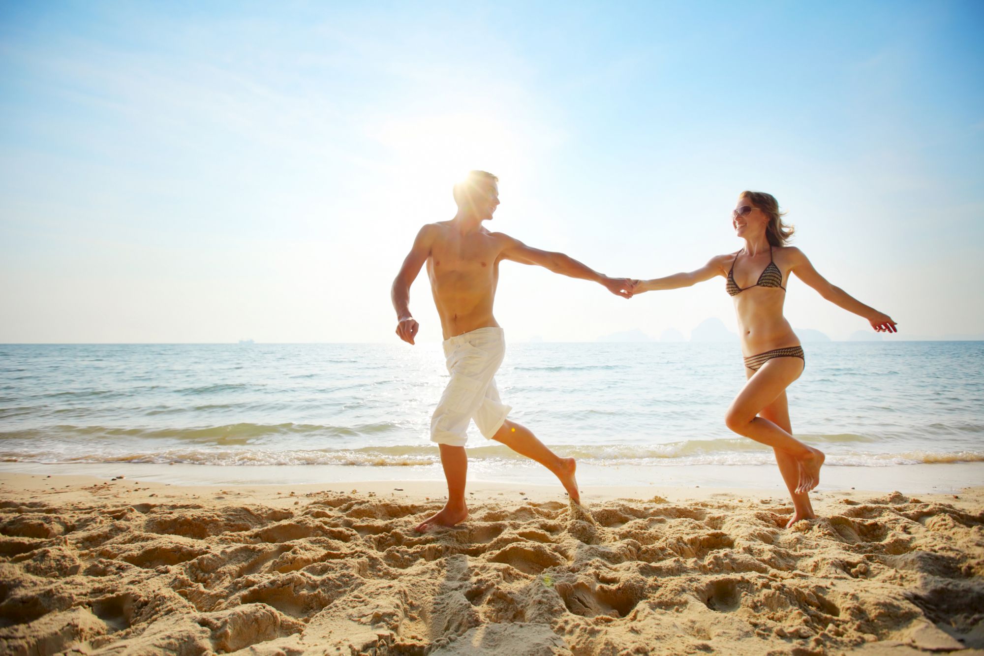 A couple holding hands, running and smiling on a sunny beach, with the ocean waves in the background.