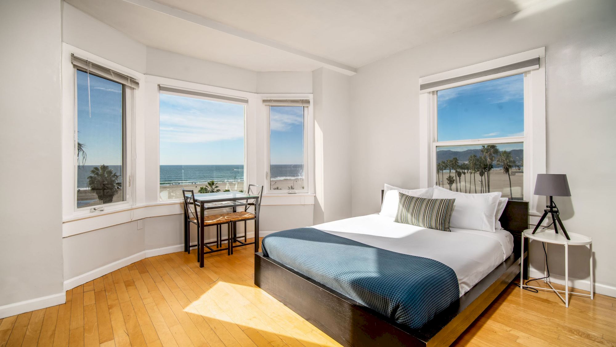 A bright bedroom with a large bed, side table, lamp, and small desk by bay windows offering a scenic ocean view under a clear blue sky.