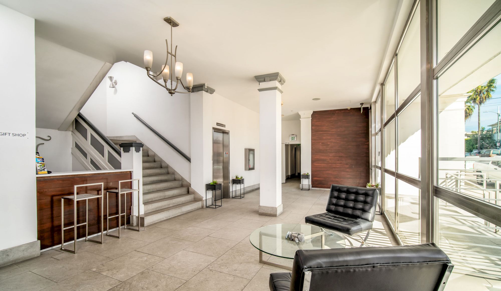 The image depicts a modern lobby area with a seating area, glass table, staircase, bar counter with stools, and large windows letting in natural light.