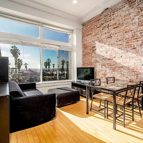 A modern living room with a large window, exposed brick wall, black furniture, a TV, and a view of palm trees and the ocean outside.