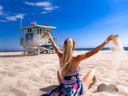 A person with long hair sits on a sandy beach, arms raised, with a colorful dress, near a lifeguard tower under a clear blue sky.