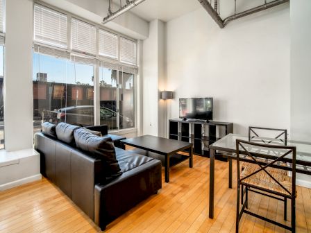 A modern living room with a black couch, coffee table, TV on a stand, glass dining table with chairs, and large windows with blinds ending the sentence.